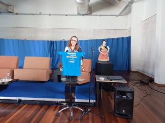 Presenter Liliana Fernandes at a high desk in front of a stage with chairs